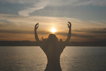 Silhouette of young woman which is looking at a sky in a sunset rays and is stretching her hands to a sun on water lake background.