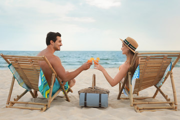 Canvas Print - Happy young couple with cocktails sitting on deck chairs at sea beach