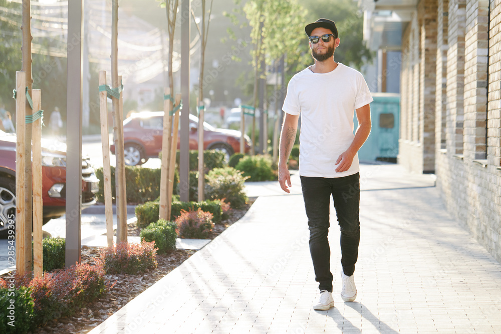 Hipster handsome male model with beard wearing white blank t-shirt and a baseball cap with space for your logo or design in casual urban style - obrazy, fototapety, plakaty 