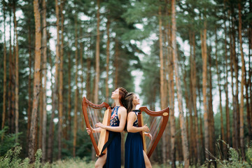 Two women harpists stand at forest and play harps against a background of pines.