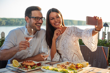 smiling happy young couple posing for a selfie, eating dinner in a riverside restaurant. husband and