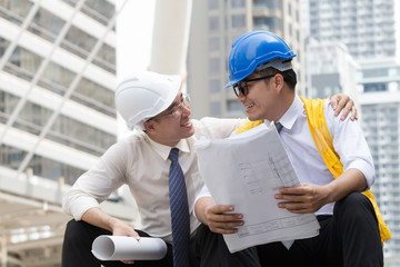 Asian engineer man and worker in uniform are happy with successful construction project and work plan. They are holding blueprint paper and wear safety helmet and yellow vest wiht smiling face.