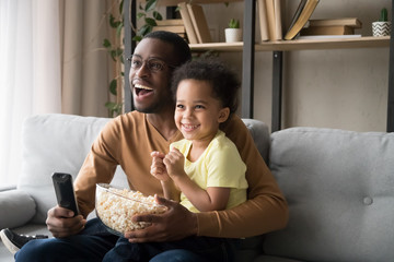 Wall Mural - Happy African American father with son watching tv, eating popcorn