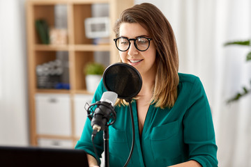 Wall Mural - technology, mass media and people concept - woman in glasses with microphone and laptop computer talking and recording podcast at studio