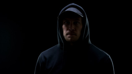 Confident male gangster in hoodie looking at camera against black background