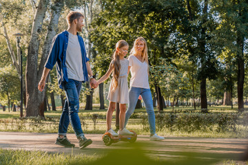 Side view of young smiling parents walking in park with their cute little girl riding hoveboard