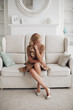 © Вячеслав Косько - Gorgeous pretty lady sitting in luxury apartment and holding hands on knees. Beautiful young woman in elegant beige dress posing on white sofa. Happy blonde model relaxing after long working day.