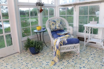 Beautifully decorated interior of a greenhouse/summer house with stenciled floor, white wicker chaise lounge and table, with blue and yellow floral cushions and plaid throw and blue planters