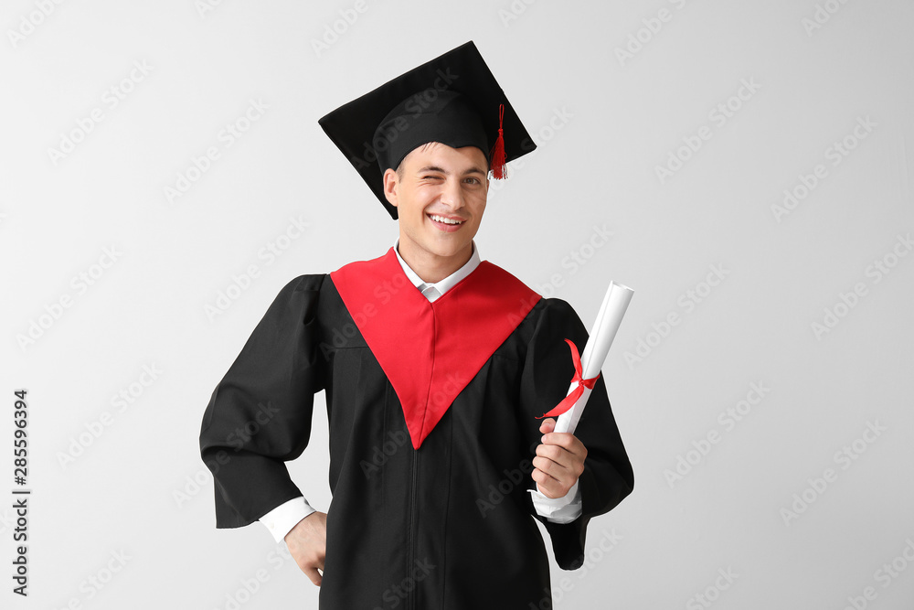 Young male student in bachelor robe and with diploma on light background