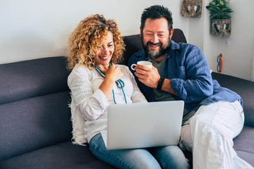 Happy adult aged couple caucasian people at home with personal laptop computer and internet connection - together in indoor leisure activity with technology sitting on the sofa and smiling