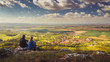 © Roman - Two people (couple - man and woman) sitting on a mountain on a stone and looking into the valley on beautiful autumn landscape with village, vineyards and fields. Blue sky, white clouds. Palava, Czech