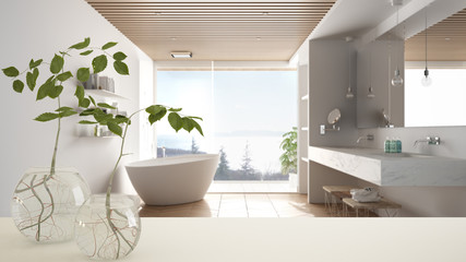 White table top or shelf with glass vase with hydroponic plant, ornament, root of plant in water, branch in vase, house plant, bathroom with bathtub in the background, interior design