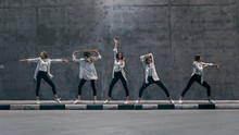 Multiple Exposure Shot Of A Cool Cloned Young Blond Woman With Long Hair Poses In Three Dance Poses On A Street Next To A Big Concrete Wall. She's Wearing A Striped White Shirt And Dark Jeans.