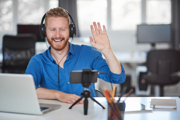 Hardworking employee having video call over smart phone while sitting in office and waving. On head are headphones and on desk laptop. Start up business concept.