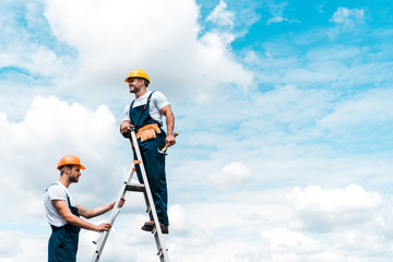 Wall Mural - cheerful repairmen standing on ladder and smiling against blue sky with clouds