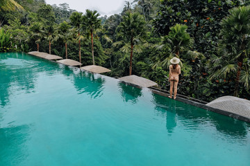 Infinity pool at luxurious exotic island. Back view of woman stay on edge of pool and enjoy jungle view wearing beige bikini and hat. Vacation concept