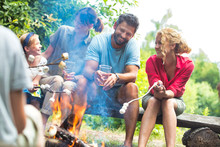 Happy Family Sitting With Woman While Roasting Marshmallows Over Burning Campfire At Park