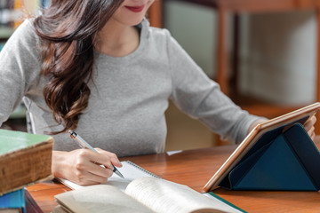 Wall Mural - Closeup Asian young Student hand writing homework and using technology tablet in library of university or colleage with various book and stationary over the book shelf background, Back to school