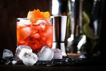 Americano Alcohol cocktail with red vermouth, bitter, soda, orange zest and ice, dark wooden bar counter background, bar tools, selective focus