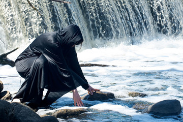 Hooded Woman in black robes in front of the Waterfall. Witches. Halloween and Gothic concept. Witchcraft and magic.