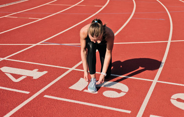 Wall Mural - Young sprinter woman in sportswear tying shoelaces before a race on a red-coated stadium track at sunny bright day