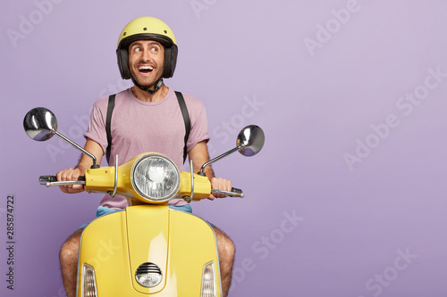 Fotografía Happy male biker or courier drives yellow scooter, wears protective helmet, casu