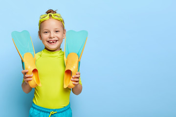 Studio shot of happy ginger small girl shows new rubber flippers, wears swim goggles, dressed in summer clothes, enjoys learning swimming, has active rest, isolated on blue wall with blank space