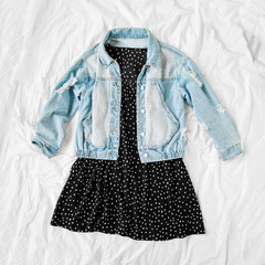 Wall Mural - Blue jean jacket and black dress on white bed. Women's stylish autumn or spring outfit. Trendy clothes. Flat lay, top view.