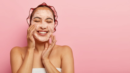 Studio shot of happy Asian lady washes face with soap, enjoys cleanliness and softness, cleans skin, closes eyes with pleasure, grins at camera, just had shower isolated on pink wall. Wellness concept