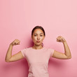 Photo of self confident serious Asian girl keeps lips folded, shows her muscle and strength, wears no make up, dressed in casual t shirt, isolated on pink wall with copy space above for information