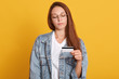 © sementsova321 - Young beautiful Caucasian woman presenting credit card for making payment, adorable lady looks at card, wearing denim jacket and white t shirt, thinks about new purchase, has dark straight hair.