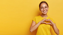 Lovely Korean Woman With Happy Smile, Shapes Heart With Both Hands, Expresses Love To You, Wears Round Spectacles And Yellow T Shirt, Says Be My Valentine Flirts With Boyfriend Poses Indoor Copy Space