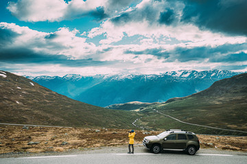 Aurlandsfjellet, Norway. Young Caucasian Woman Lady Tourist Traveler Photographer Taking Pictures Photos Near Parked Car. Aurlandsfjellet Scenic Route Road In Summer Norwegian Countryside Landscape