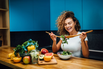Wall Mural - Young and happy woman eating healthy salad sitting on the table with green fresh ingredients indoors . Healthy life.