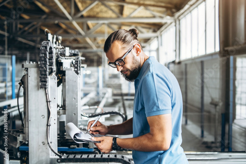 Fotomural Young engeneer manager with beard checking manufactory, workplace and machinery on big factory