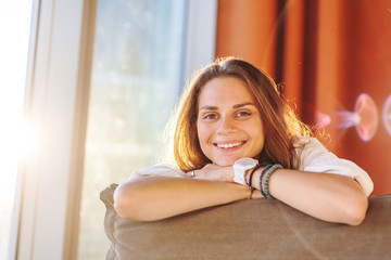 Beautiful young woman with a happy smile looks at the camera, sits in the apartment in the sun from the window