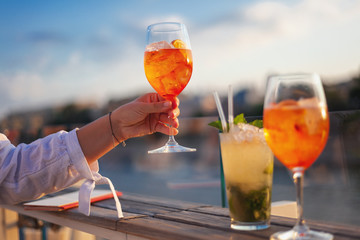 Still life shot of woman hand holding summer aperol italian cocktail, city sunset background