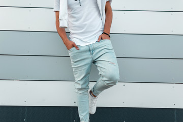 Fashionable young man in blue trendy jeans in a stylish white t-shirt in leather shoes posing near a modern building in the city. New collection of men's summer clothes. Youth style. Close-up.