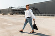 © alones - Trendy young man hipster in stylish white and denim clothes in vintage sandals with a black cloth bag walks outdoors on a sunny day. Modern guy in sunglasses in the city. Summer fashionable menswear.