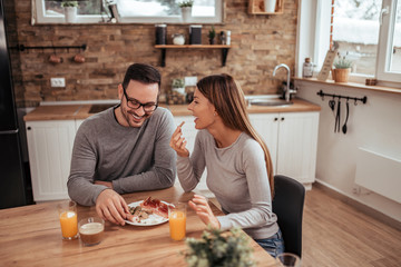 Wall Mural - Happy young spouses eating breakfast in modern kitchen with wooden table and brown stone wall.