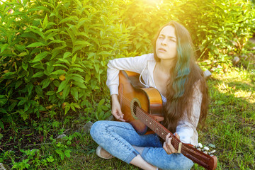 Young hipster woman sitting in grass and playing guitar on park or garden background. Teen girl learning to play song and writing music. Hobby, lifestyle, relax, Instrument, leisure, education concept