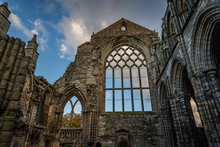 The Ruins Of Holyrood Abbey Still Stand Tall Silhouetted By Blue Sky And Fluffy Clouds Behind The Queen's Gallery Palace In Edinburgh Scotland, UK