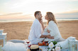 © Ulia Koltyrina - Picnic at sunset. Man and a pregnant woman sit on a bedspread, on the beach, hugging, kissing against low tide.