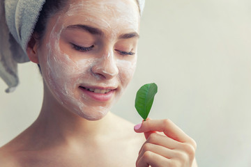 Wall Mural - Beauty portrait of woman in towel on head with white nourishing mask or creme on face and green leaf in hand, white background isolated. Skincare cleansing eco organic cosmetic spa concept