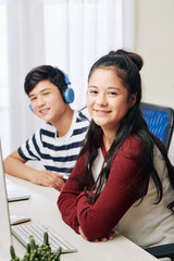 Wall Mural - Smiling mixed-race pretty teenage girl sitting in front of computer at her desk, her cheerful brother listening to music in background