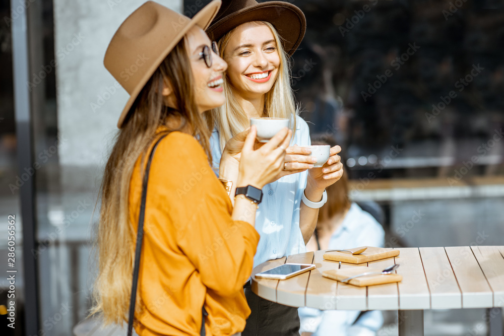 Two female best friends spending time together on the cafe terrace, feeling happy, enjoying coffee during a summer day - obrazy, fototapety, plakaty 