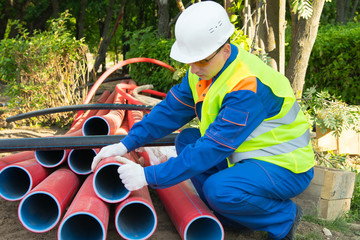 Wall Mural - builder, in a white helmet, pulls out red plastic pipes into a trench, for laying an electric cable through it, close-up