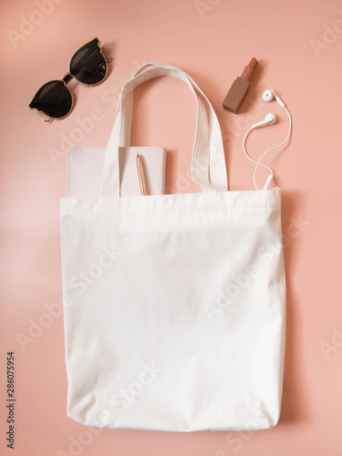 Mock up design bag concept. Top view of blank white tote bag canvas fabric w/ sunglasses, lipstick, notebook, pen and headphone on coral background. Empty eco shopping bag. Flat layout. Vertical.