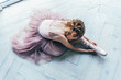 Young classical ballet dancer girl in dance class. Beautiful graceful ballerina in pink tutu skirt puts on pointe shoes near large window in white light hall