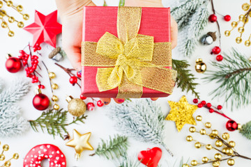 Hand holding red gift box with golden ribbon and bow against Xmas decor background. Christmas composition, flat lay top view